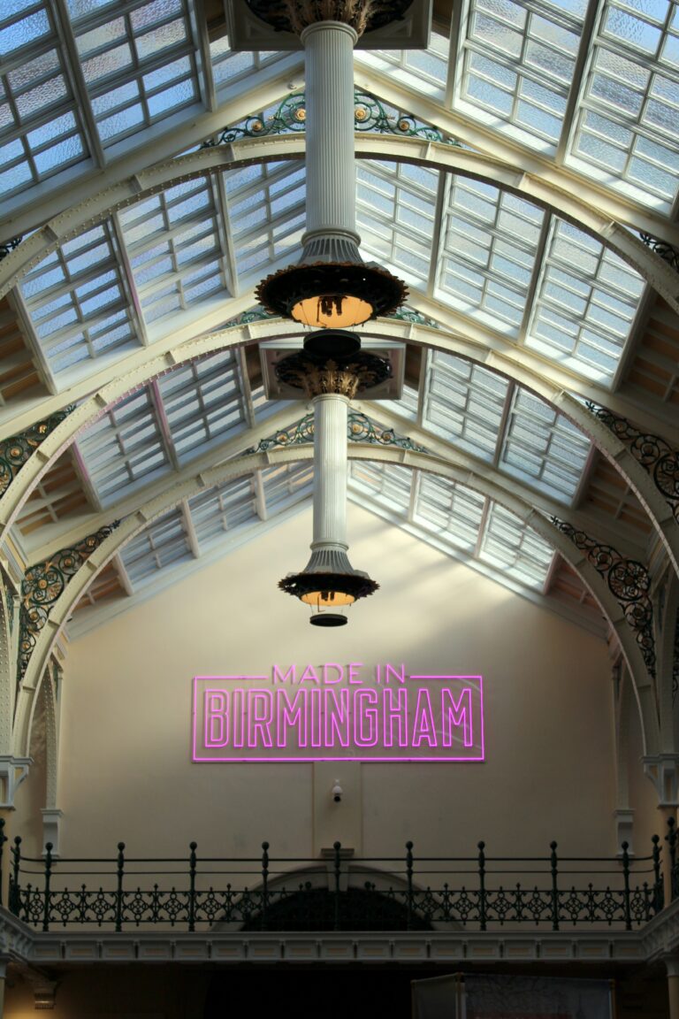A large hall with glass ceiling panels and industrial era features, and a pink neon sign saying 'Made in Birmingham'.
